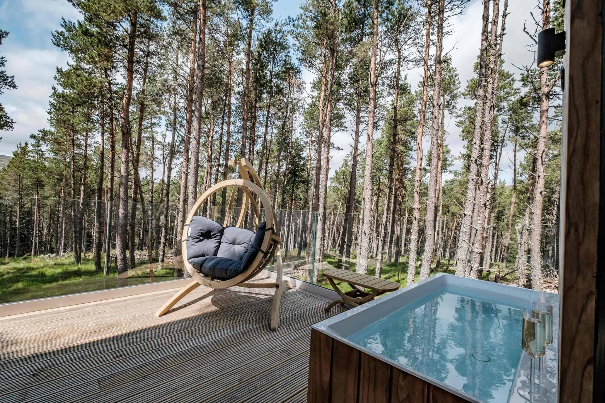 Square wood-clad outdoor bathtub on a glass-railed deck in a pine forest, with a circular hanging chair beside it