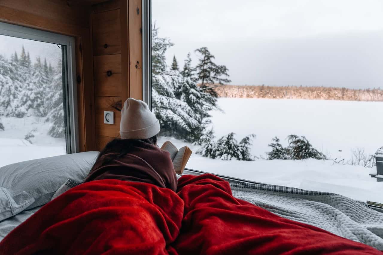 Person in a beanie reading on the Cove Cabin bed under a red blanket, snow-covered lake and evergreens through the window