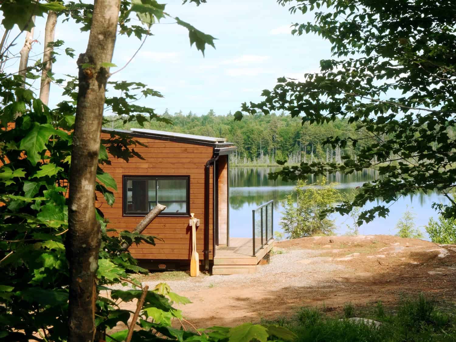 Wood-sided Cove Cabin on a rocky lakefront clearing, glass-rail deck, canoe paddle by the door, summer trees and still water