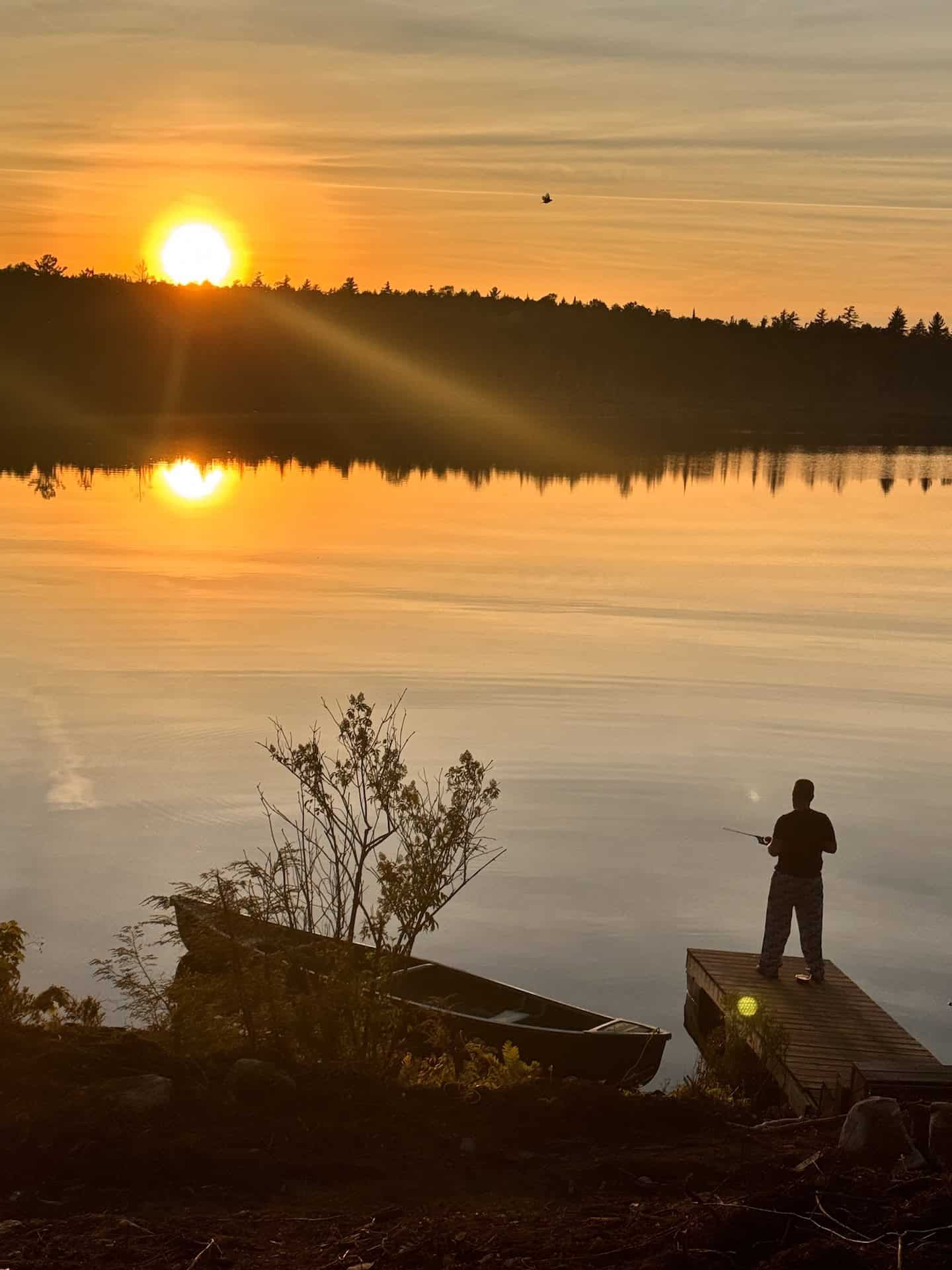 Sunset over the lake: a person fishing from a wood dock, canoe on the rocky shore, orange sun over the far trees