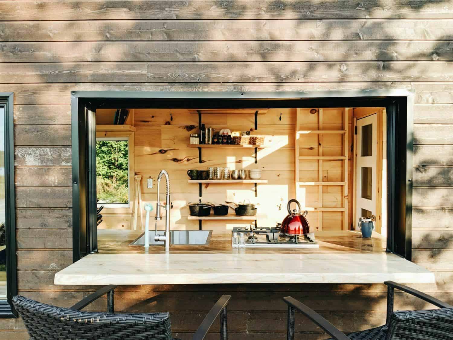 Cove Cabin pass-through summer bar: live-edge wood slab through a black-framed opening, two black chairs outside, sink and two-burner stove inside