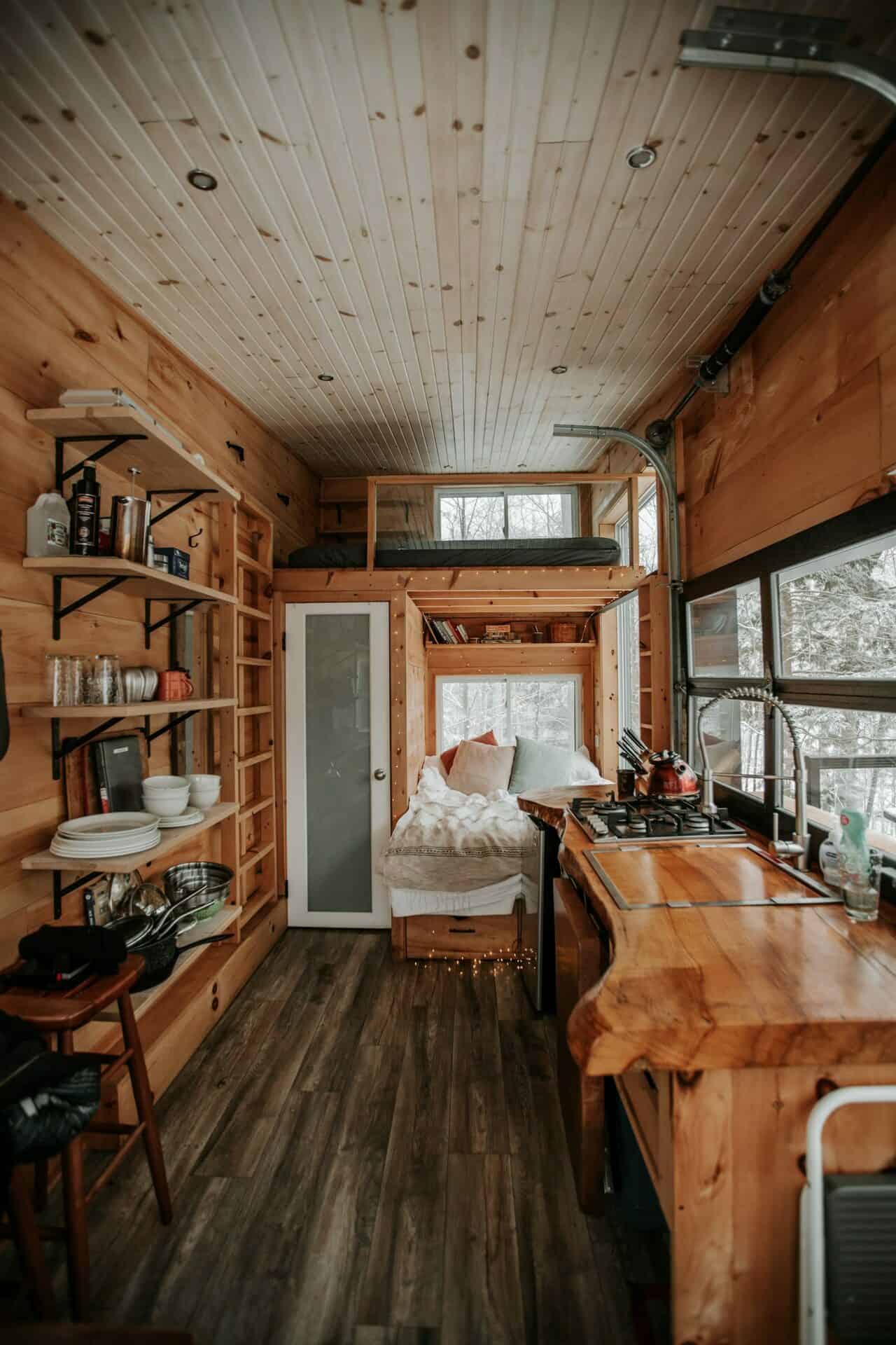 Cove Cabin interior looking toward the bed nook: live-edge counter with two-burner stove and red kettle, open shelves, loft ladder, winter woods through the glass wall