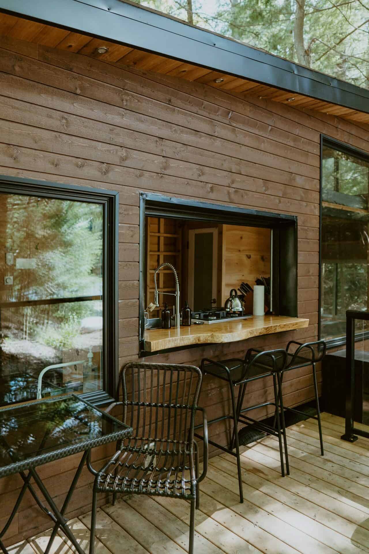 Cove Cabin deck in summer: live-edge pass-through bar with two black stools, black bistro table at the window, forest in the glass