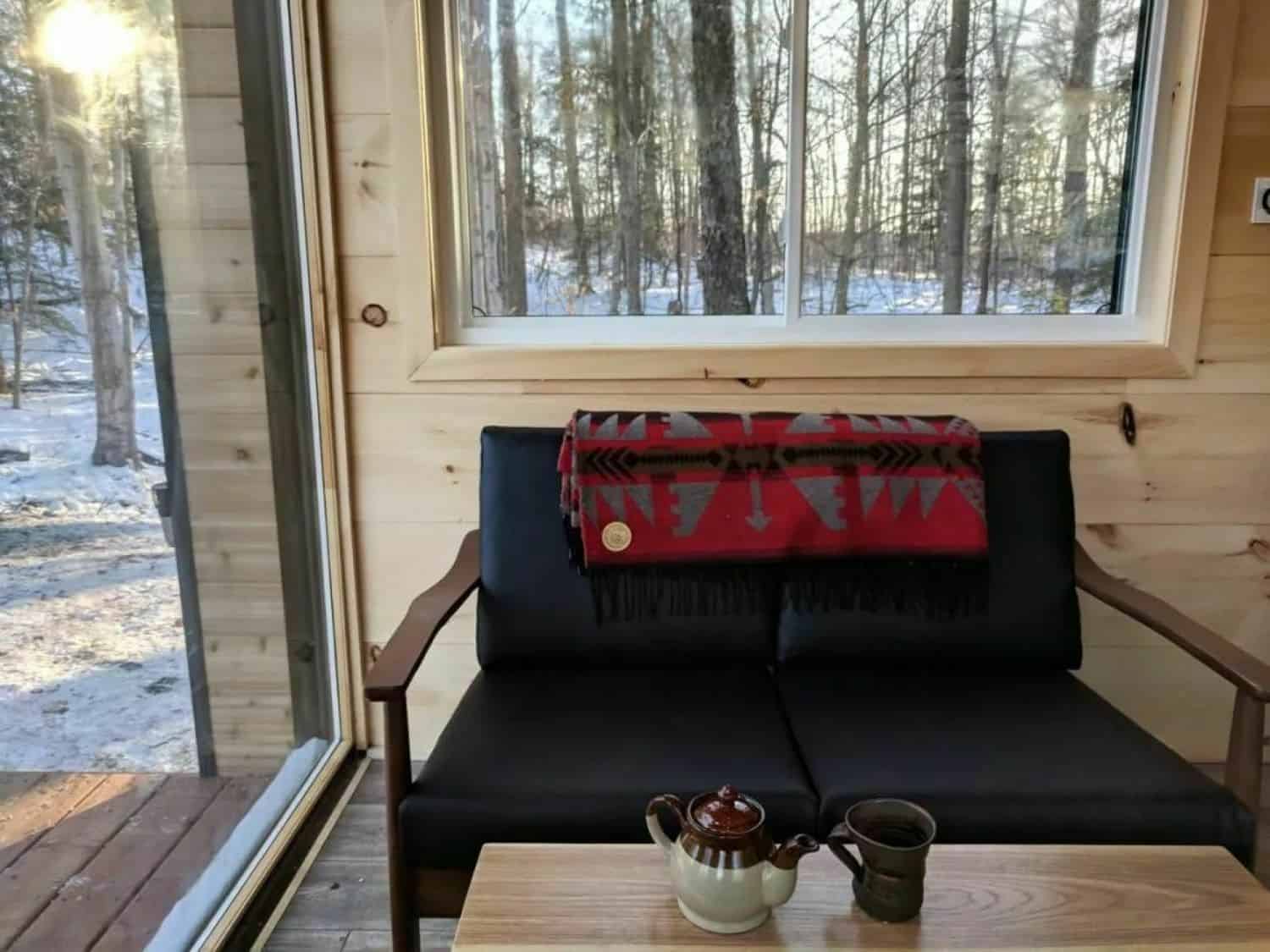Black love seat with a red geometric blanket, wood coffee table with teapot, snowy woods through the window and glass door