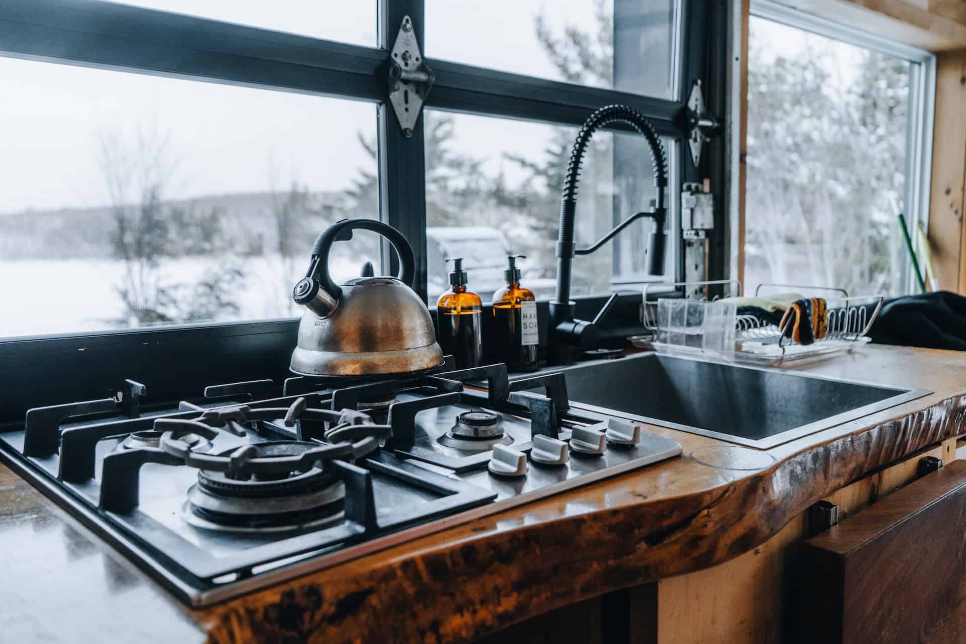 Live-edge wood counter with a two-burner gas stovetop, metal kettle, and black gooseneck faucet, snowy evergreens through the black-framed window