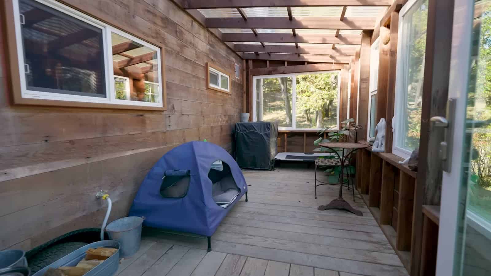 Long covered porch along the side of the tiny house with a clear roof, a dog tent bed, and a small bistro table