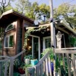 Caitlin’s dark-stained tiny house on a wooded hillside with a covered porch, sliding glass doors, and potted plants