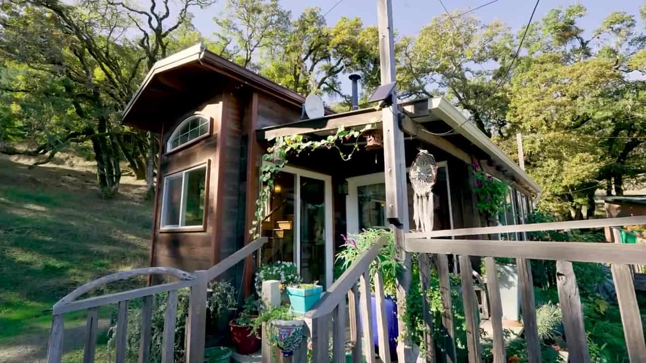 Caitlin’s dark-stained tiny house on a wooded hillside with a covered porch, sliding glass doors, and potted plants