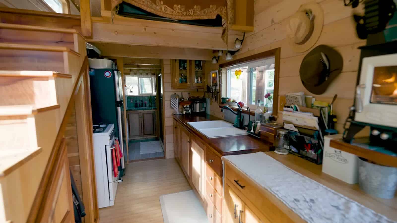 Long galley kitchen in a pine-lined tiny house with a white farmhouse sink, wood counters, compact range, and a wood stove at the far end