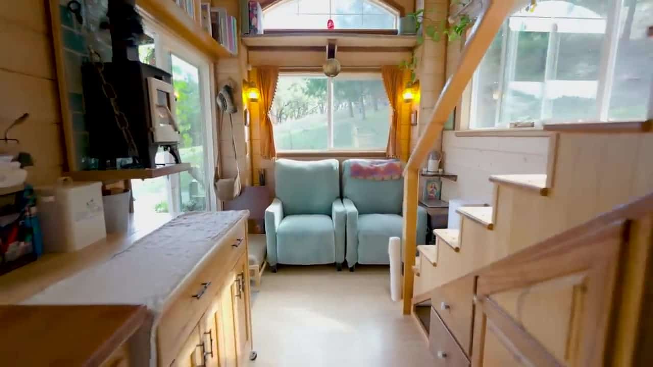 Living end of the tiny house with two light-blue recliners under a large window, wood stove to the left, and storage stairs to the right