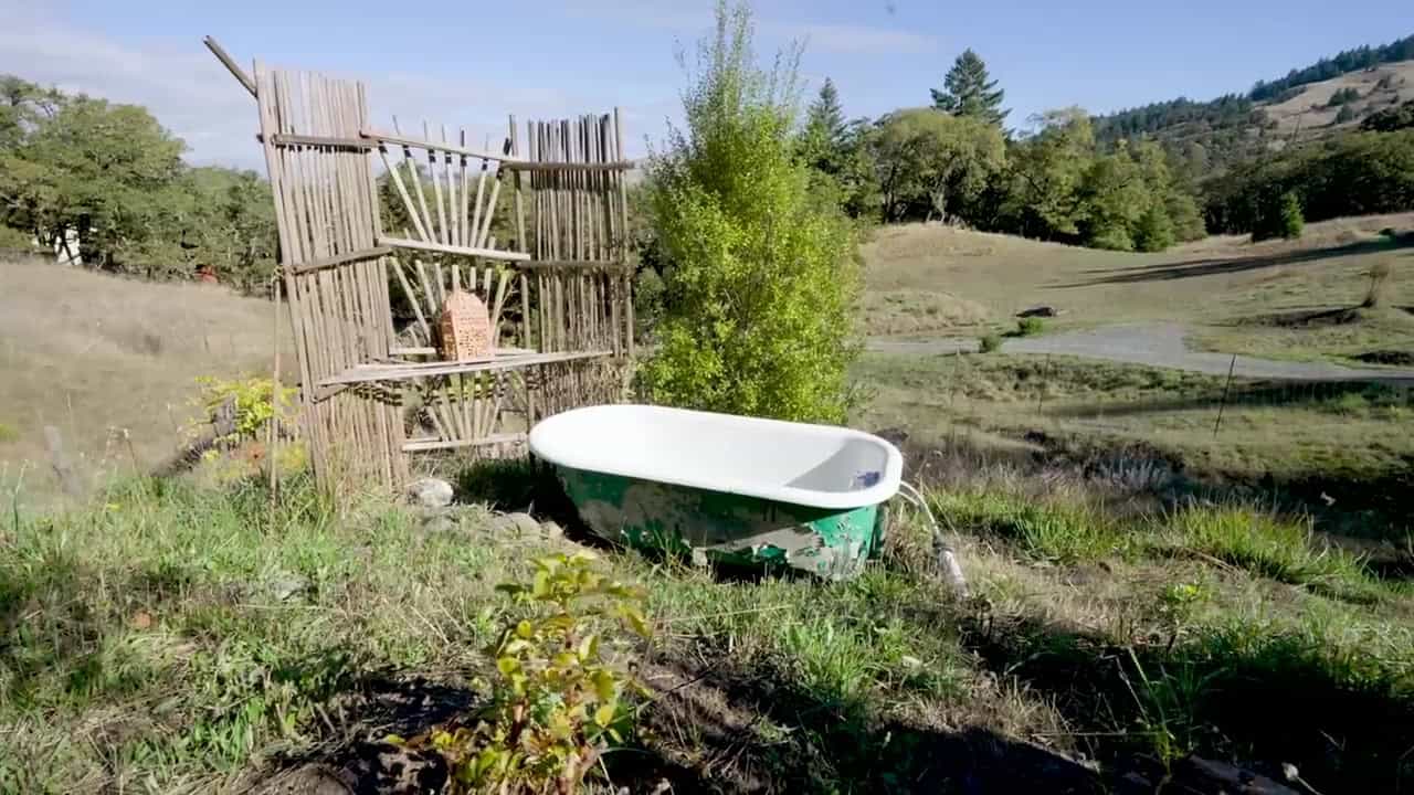 Weathered outdoor clawfoot tub on a grassy hillside beside a rustic stick privacy screen, looking out over a rolling valley