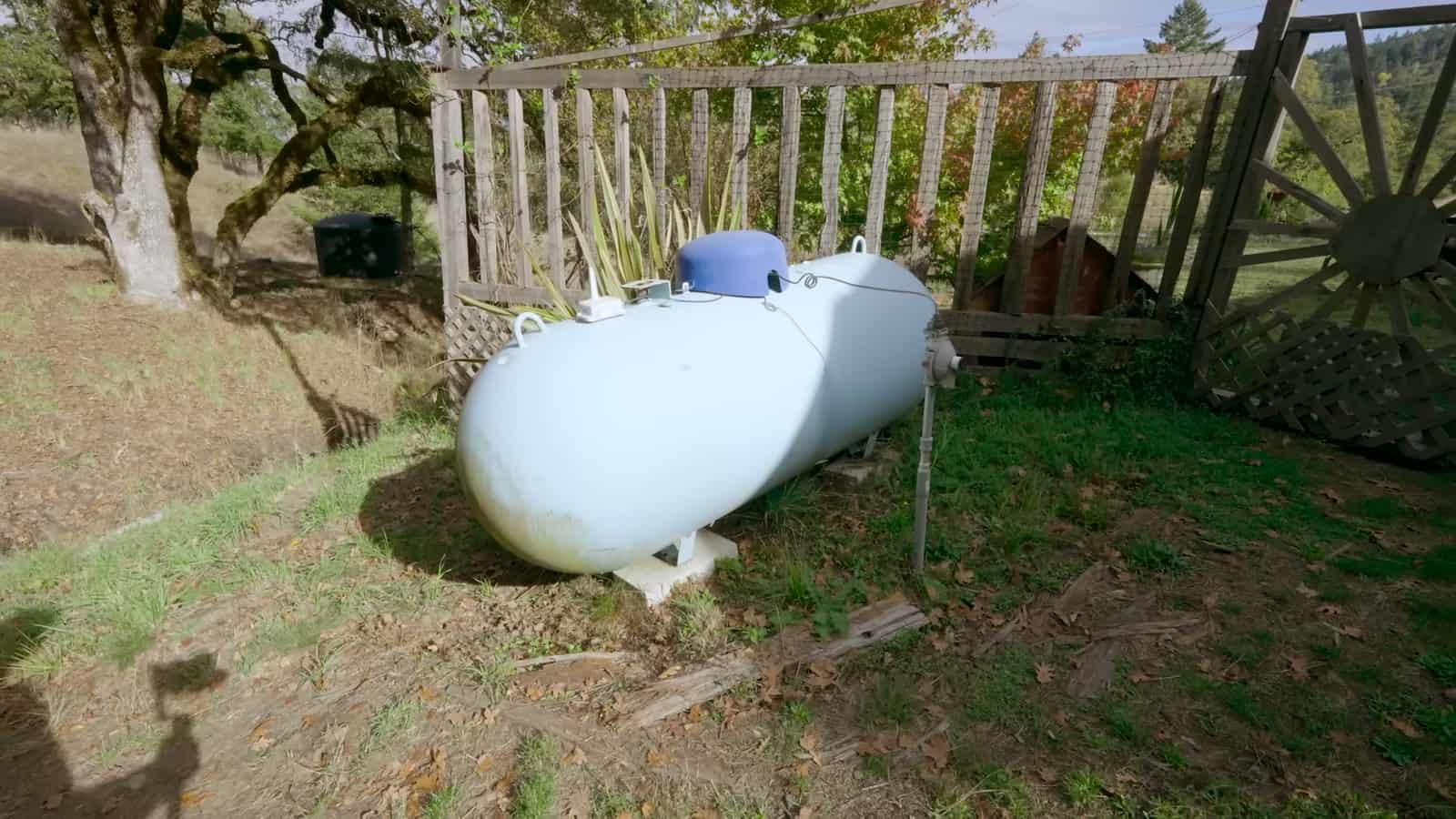 White propane tank on a grassy slope beside a weathered wooden fence with a wagon-wheel gate