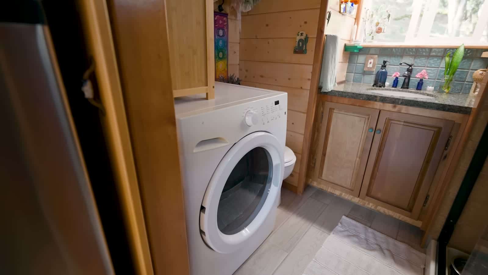 Front-load washer tucked beside a used wood vanity with a tile backsplash in the tiny house bathroom