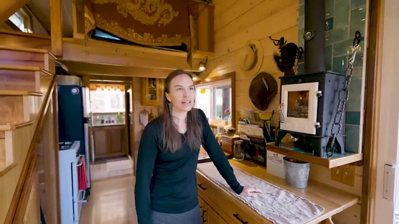 Compact black dwarf wood stove with a fire visible through the glass door and a teal tile hearth in Caitlin’s tiny house