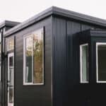 Dark California Tiny House Vesta 32 with white vinyl windows, a white-framed glass door and transom in the middle section, standing-seam metal on the end walls, and a white mini-split condenser at the far corner