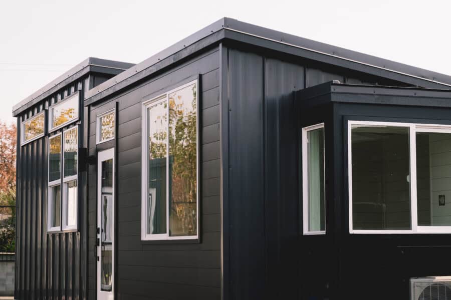 Dark California Tiny House Vesta 32 with white vinyl windows, a white-framed glass door and transom in the middle section, standing-seam metal on the end walls, and a white mini-split condenser at the far corner