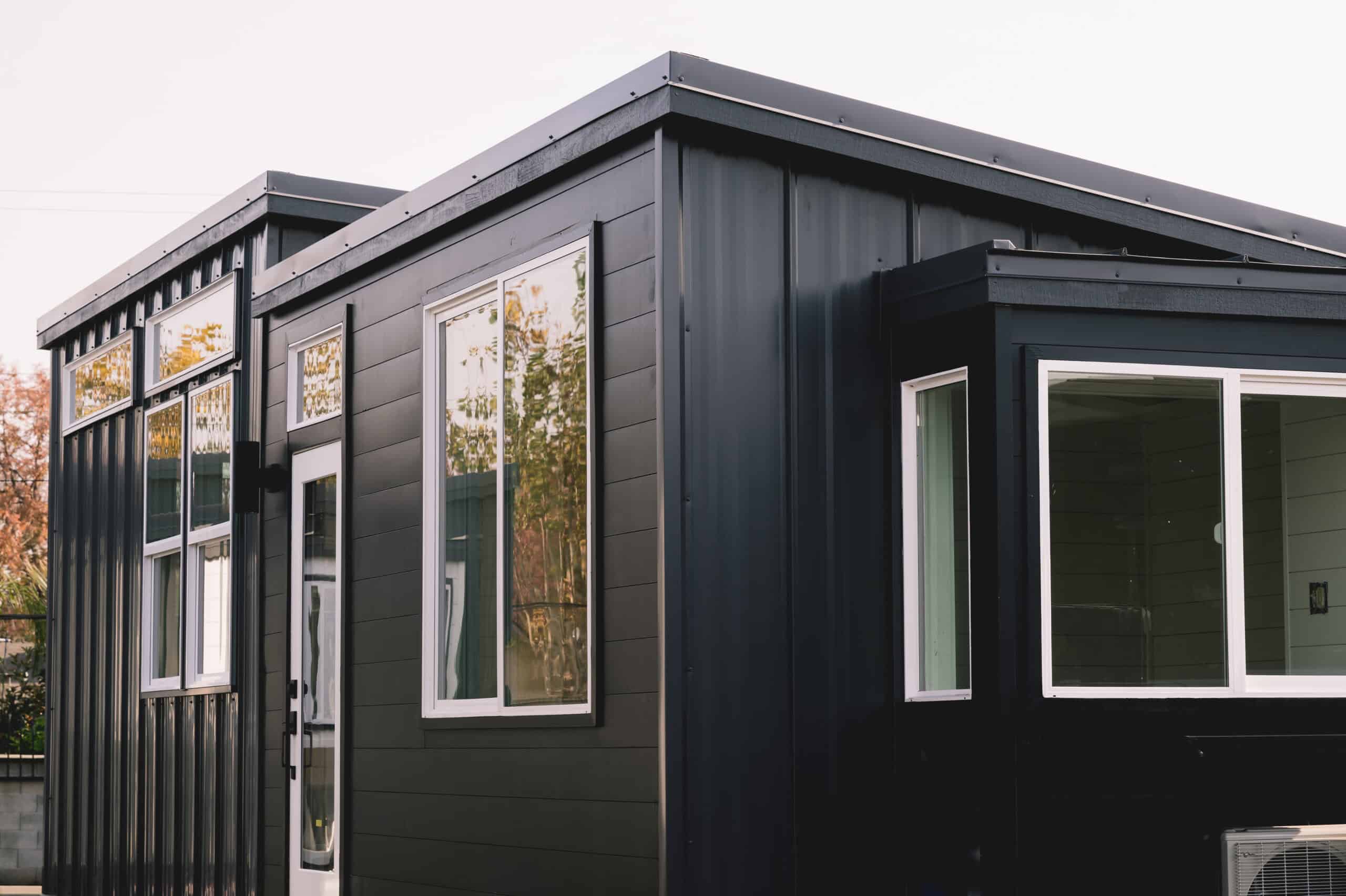 Dark California Tiny House Vesta 32 with white vinyl windows, a white-framed glass door and transom in the middle section, standing-seam metal on the end walls, and a white mini-split condenser at the far corner