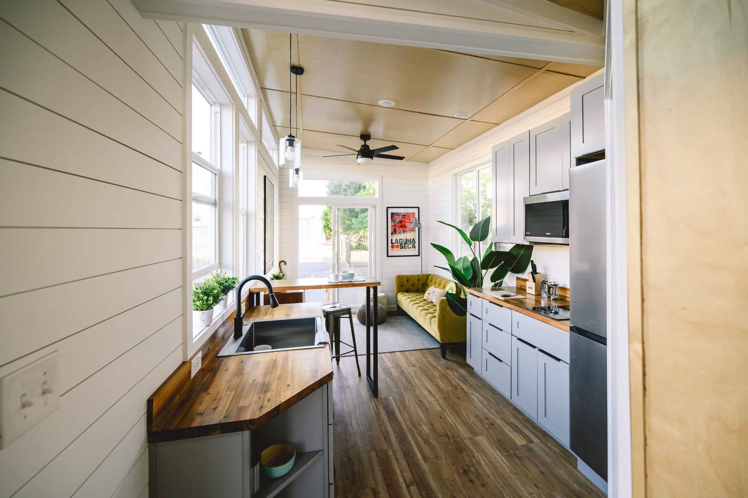 Looking down the Vesta 32 galley: grey cabinets with black hardware, acacia butcher block, two-burner induction, microwave, apartment-sized fridge, high bar with two stools, yellow sofa, sliding glass at the far end, black ceiling fan