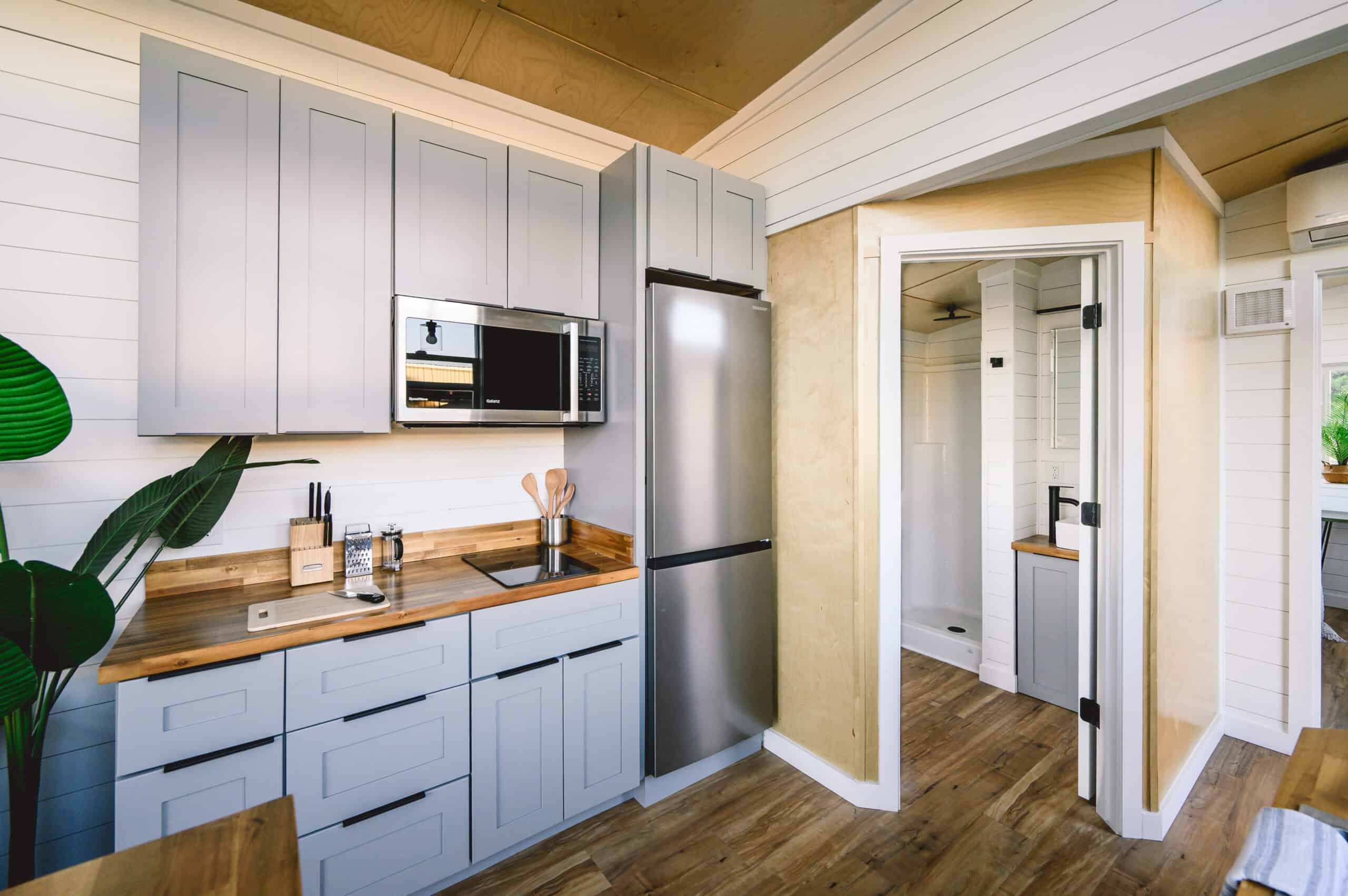 Vesta 32 kitchen close: two-burner induction in acacia butcher block, under-cabinet microwave, apartment-sized fridge in a nook, grey cabinets; doorway shows a white shower insert and a grey vanity