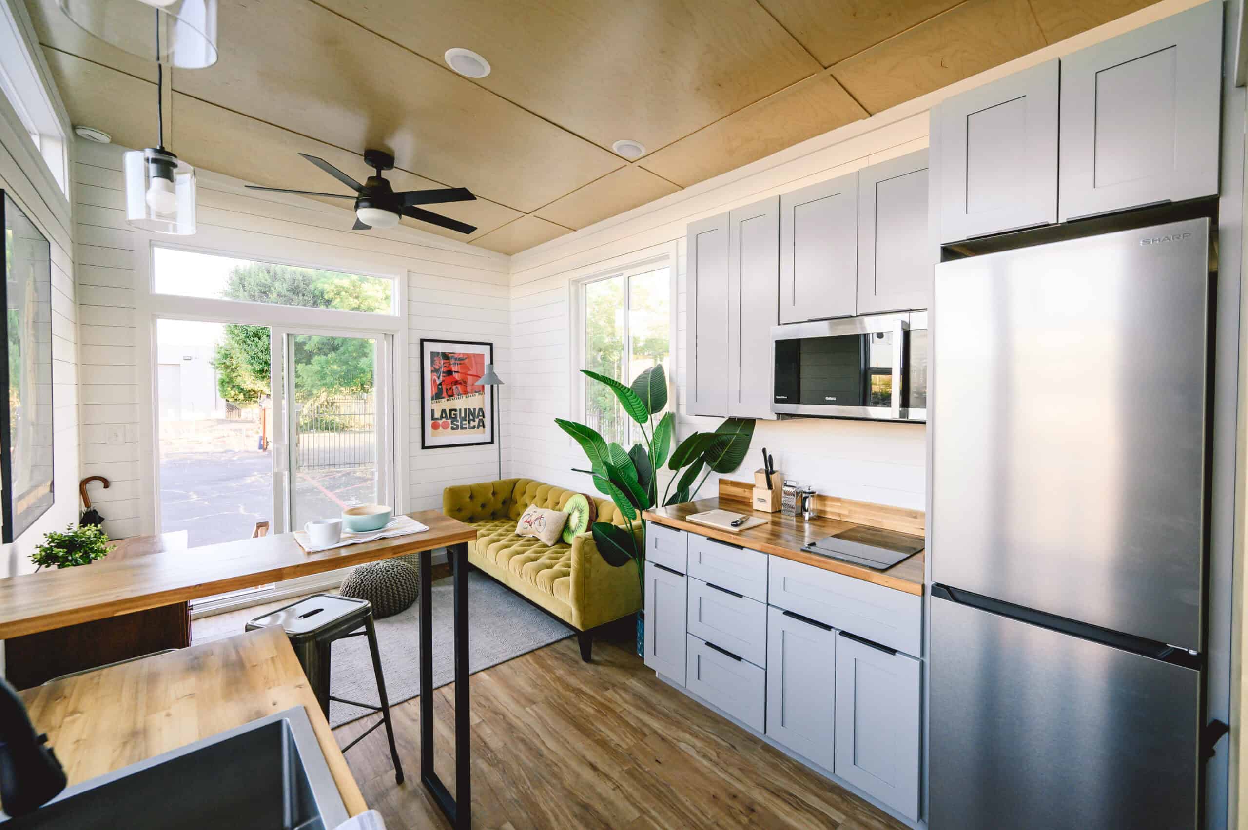 Vesta 32 from the sink toward the living room: grey cabinets, acacia butcher block and bar, apartment-sized fridge, mustard sofa, sliding glass door, black ceiling fan, painted birch walls and ceiling