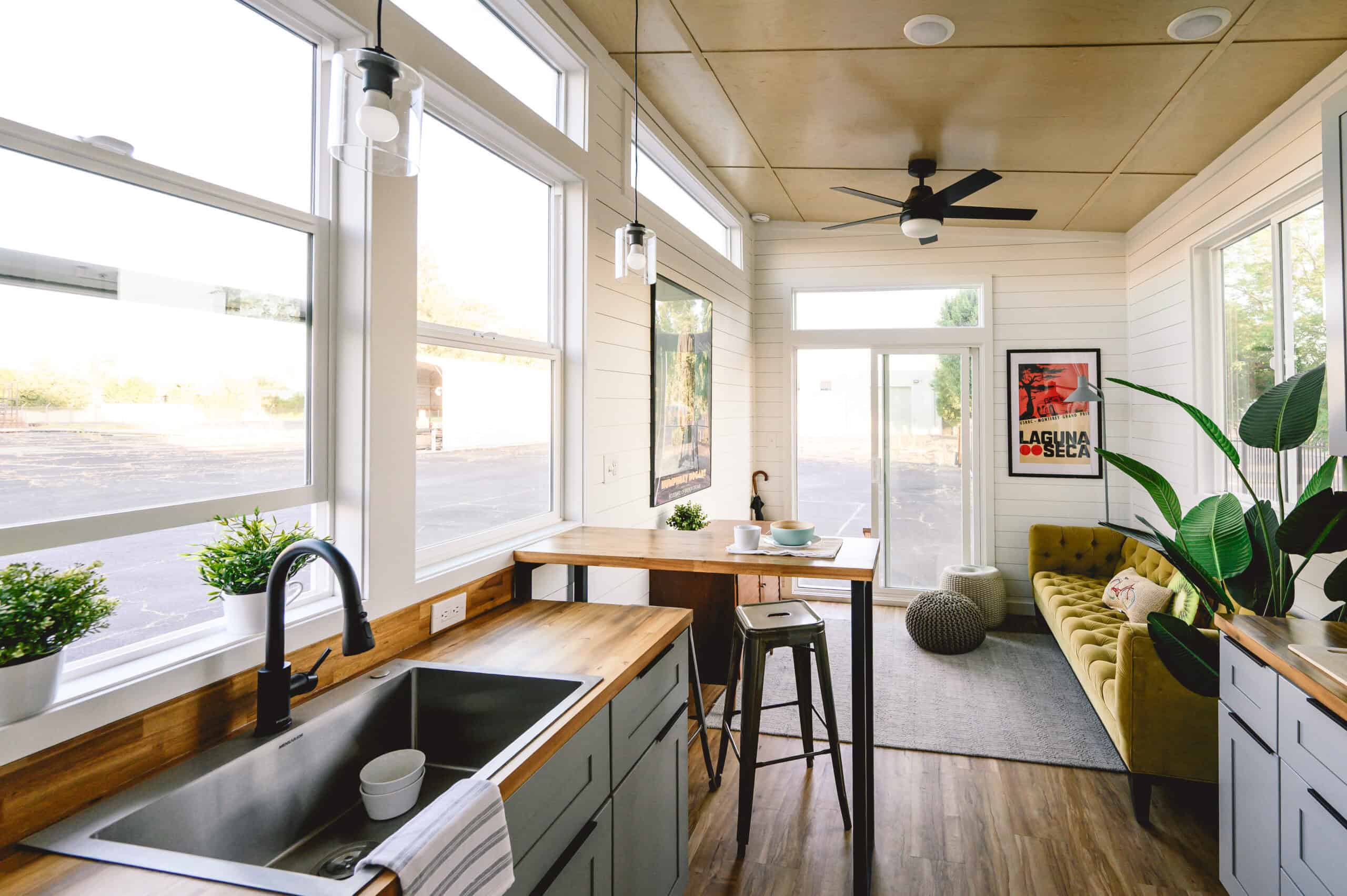 Vesta 32 walkway: stainless sink and black faucet under white vinyl windows, acacia bar, yellow sofa, sliding glass at the far end, black ceiling fan, LVP floor, painted birch walls