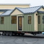 Sage-and-tan Callisto 32 tiny house on a black triple-axle deck-over trailer, standing-seam steel roof and black vinyl windows, parked at the shop