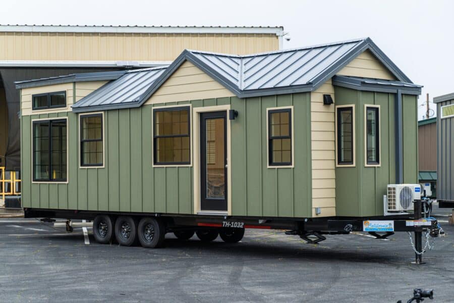 Sage-and-tan Callisto 32 tiny house on a black triple-axle deck-over trailer, standing-seam steel roof and black vinyl windows, parked at the shop