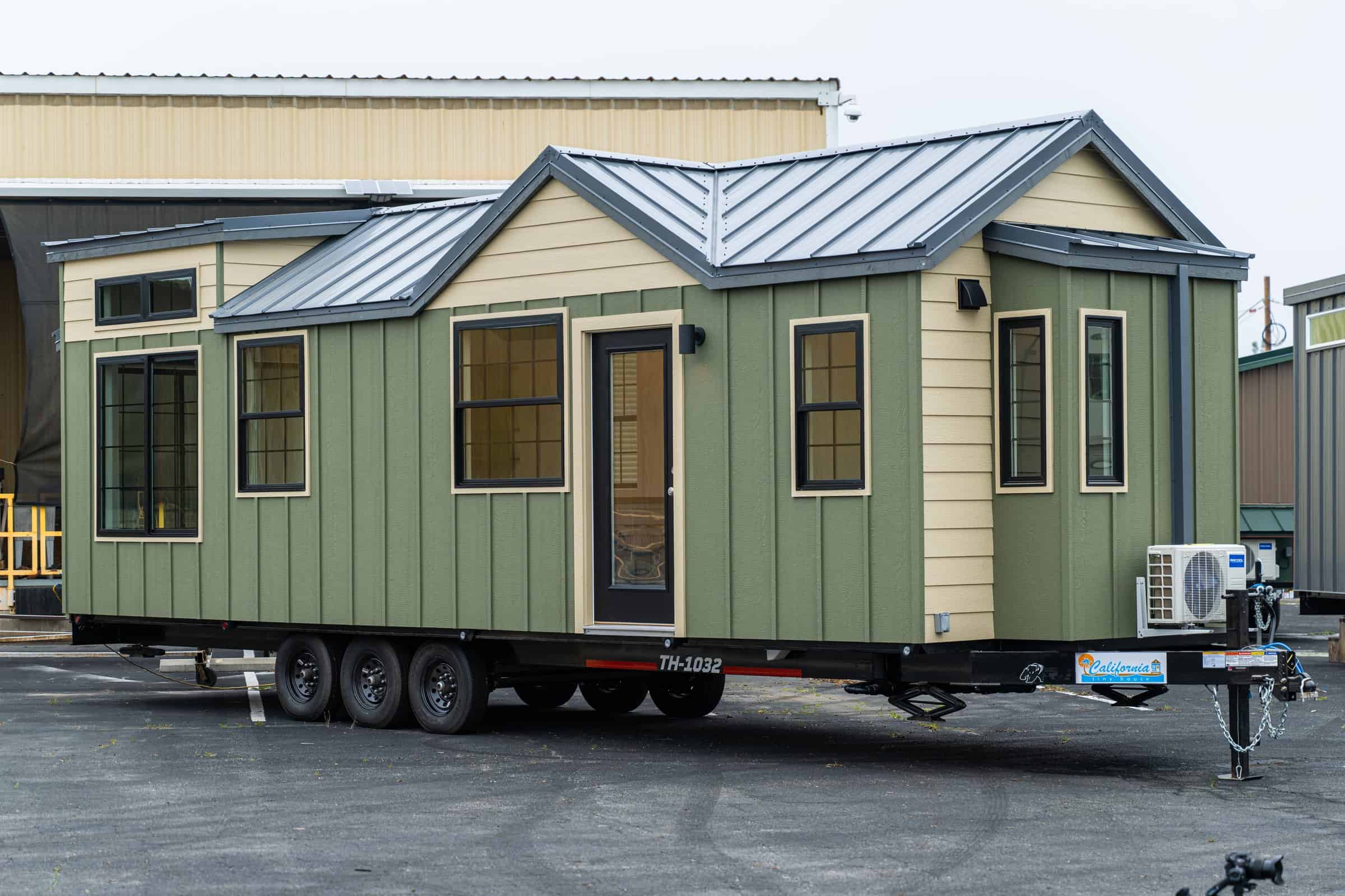 Sage-and-tan Callisto 32 tiny house on a black triple-axle deck-over trailer, standing-seam steel roof and black vinyl windows, parked at the shop