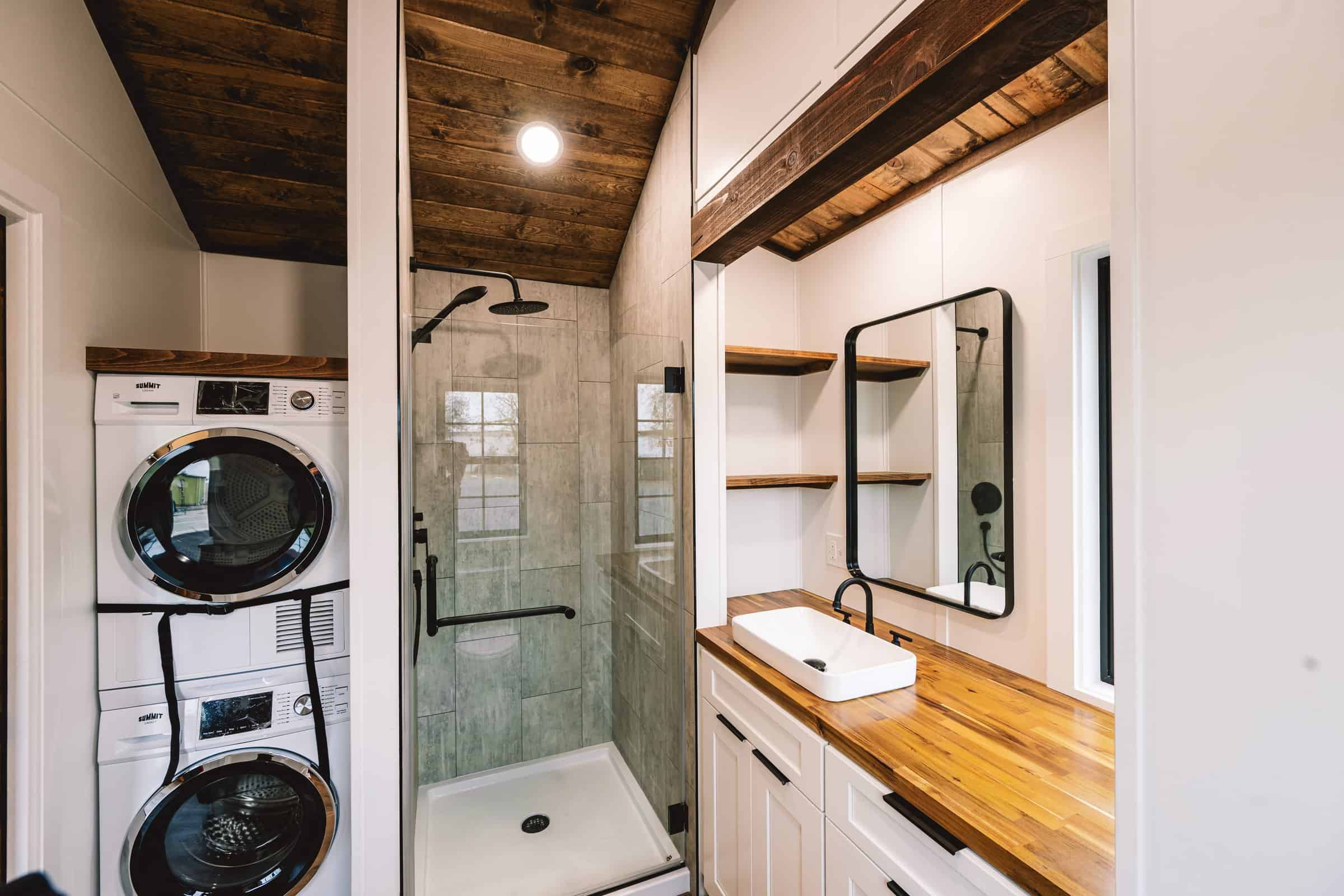 Callisto bathroom with stacked washer and dryer in a nook, glass shower with stone-look tile, butcher-block vanity, and stained wood T&G ceiling