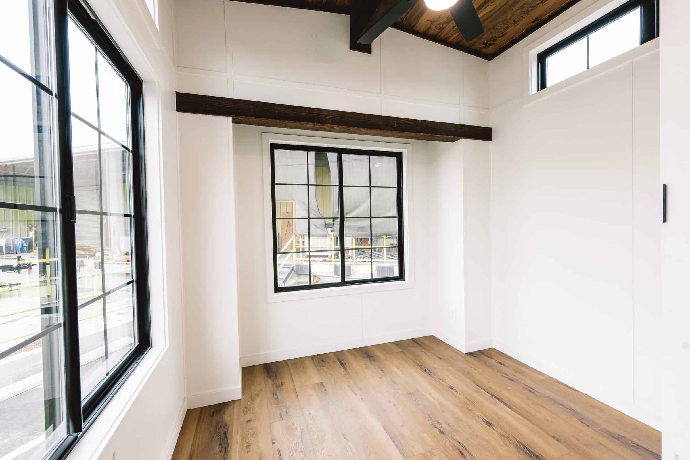 Empty single-level Callisto bedroom with stained wood T&G ceiling, open beam, black ceiling fan, painted shiplap, LVP, and large black-grid windows