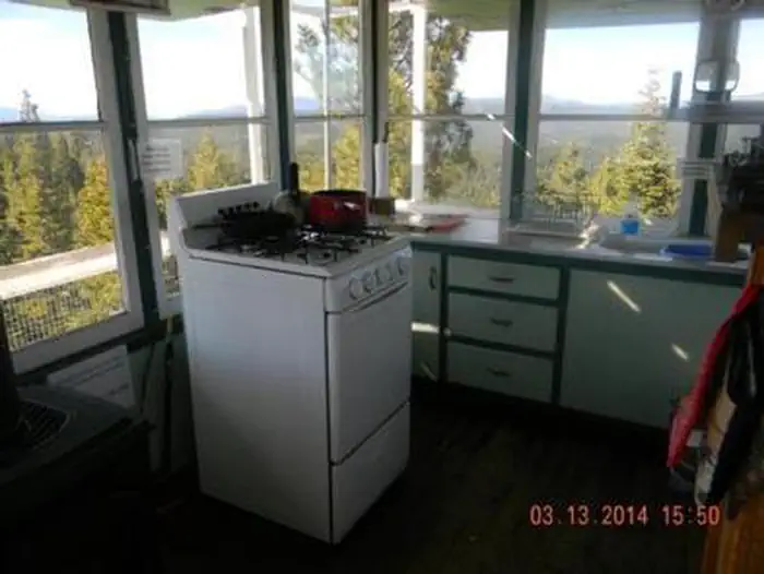 Cab interior: white four-burner propane stove and oven with a kettle and red pot, pale green cabinets, small sink, wraparound windows looking over the catwalk to pine hills