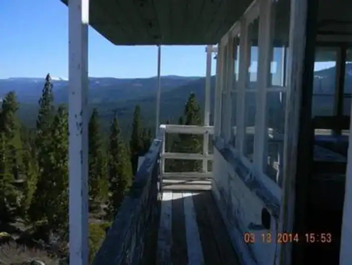 Wooden catwalk of weathered planks beside the white observation cab; multi-pane windows, white roof overhang and post, dark railing, evergreen ridges and distant mountains under a clear sky