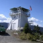 Full three-story white enclosed windmill-style lookout on a rocky ridge; external stairs wrap the tower to a windowed cab and catwalk with an American flag