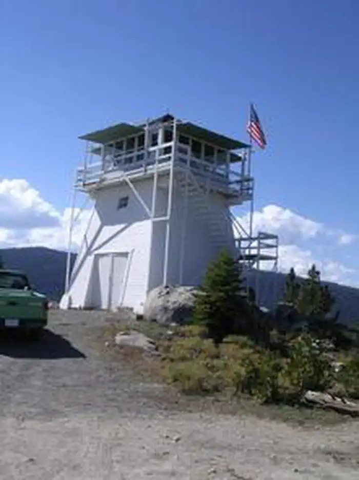 Full three-story white enclosed windmill-style lookout on a rocky ridge; external stairs wrap the tower to a windowed cab and catwalk with an American flag