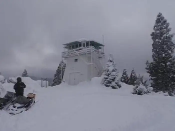 White lookout tower in deep snow, dark roof and catwalk railings snow-covered, snow-laden evergreens, overcast sky, a person in a dark winter suit near a snowmobile in the foreground