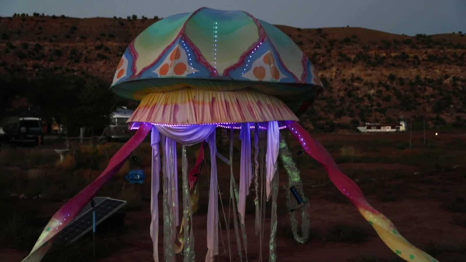 A glowing jellyfish art sculpture at dusk at Camp V with desert mesas behind it