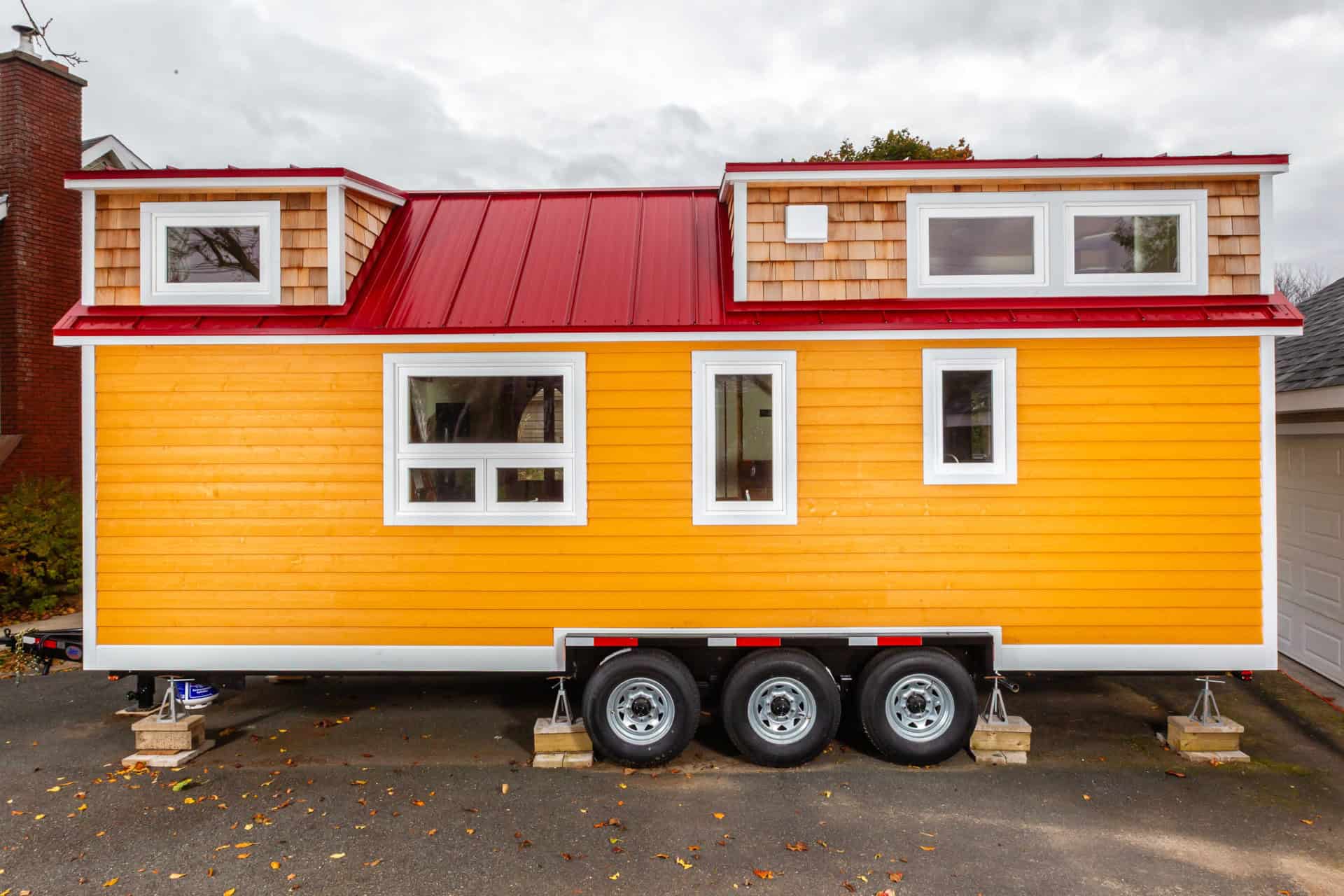 Orange tiny house with red metal roof and cedar shingle dormers on a triple axle trailer