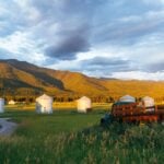 Five corrugated metal grain bins with peaked roofs in a green Kalispell meadow, forested mountains behind, an old farm truck in the foreground