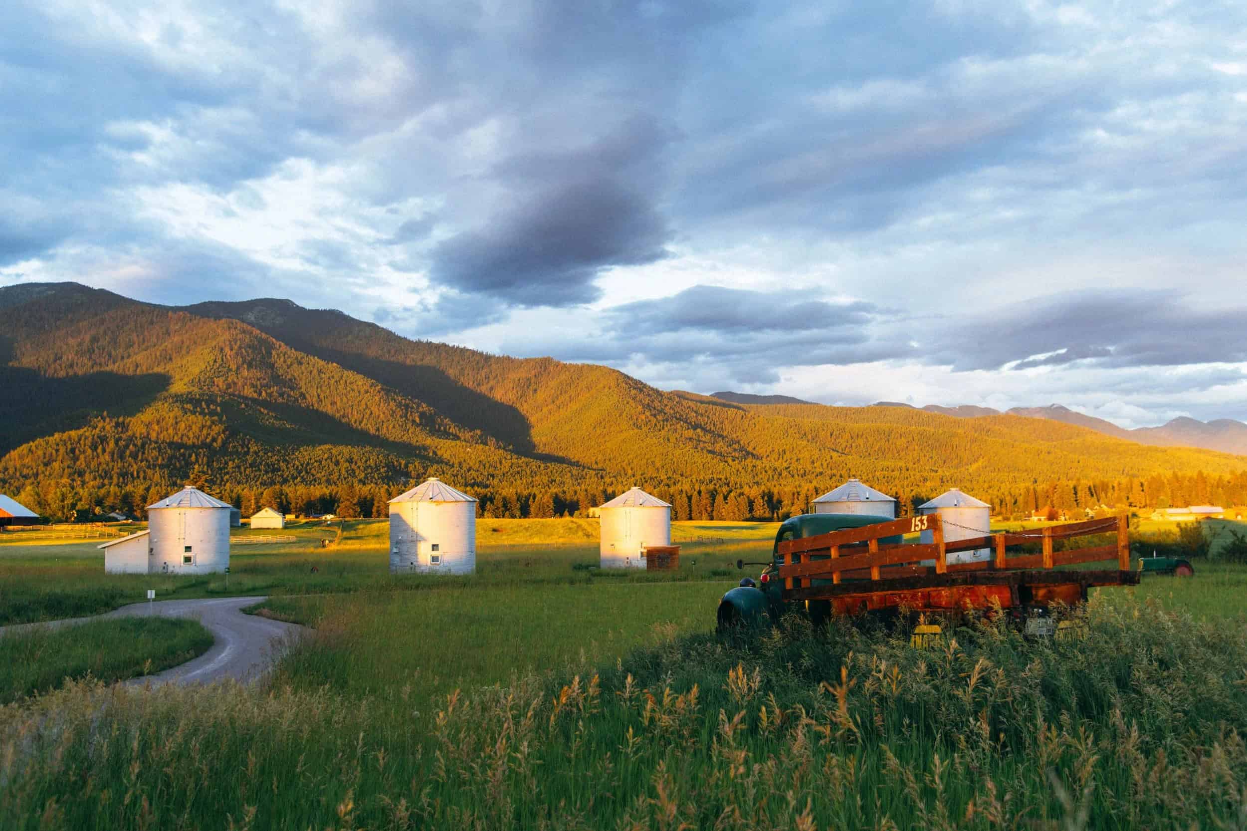 Five corrugated metal grain bins with peaked roofs in a green Kalispell meadow, forested mountains behind, an old farm truck in the foreground