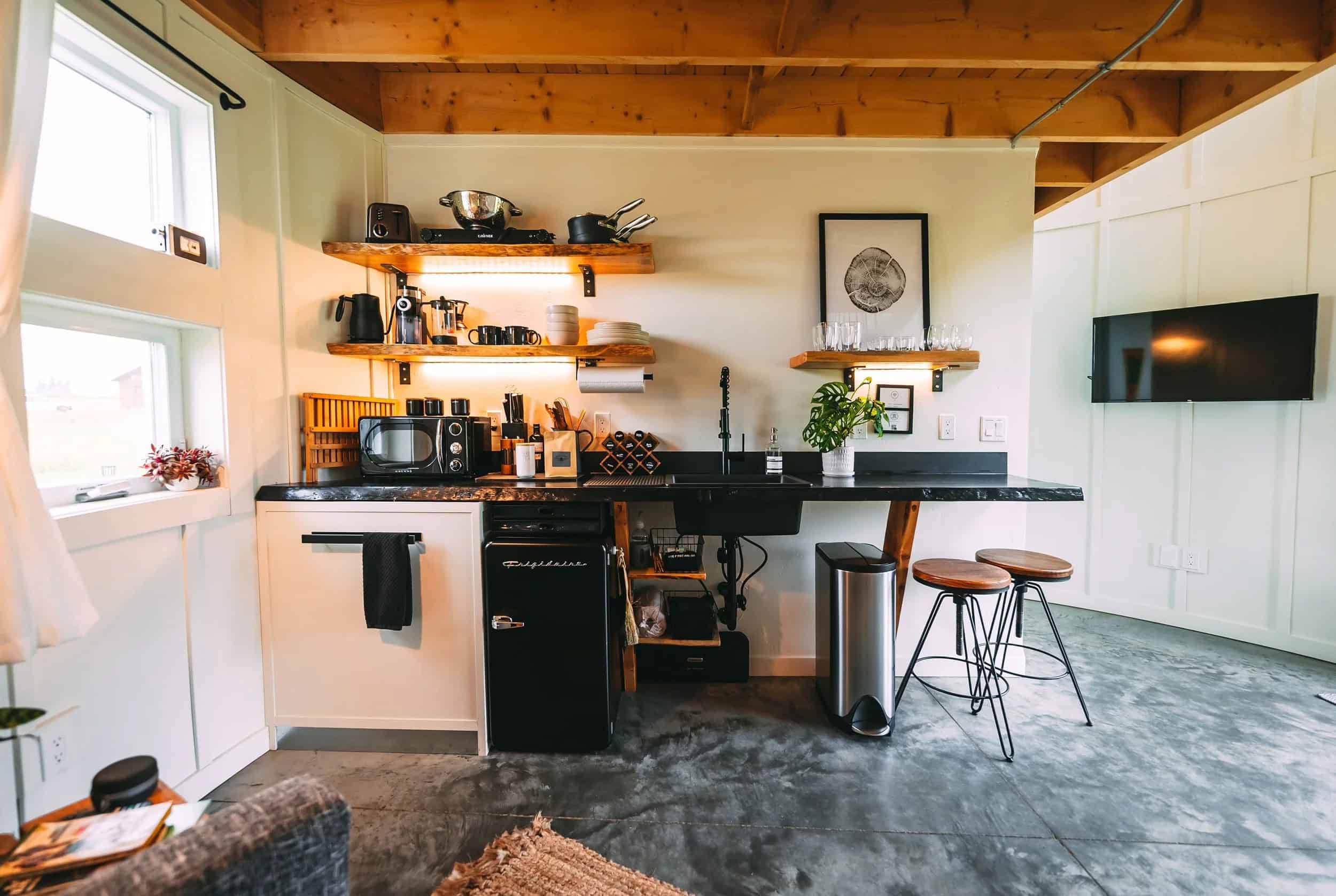 Dark kitchenette counter with a sink, compact fridge below, two wood stools, and open wood shelves on a white paneled wall