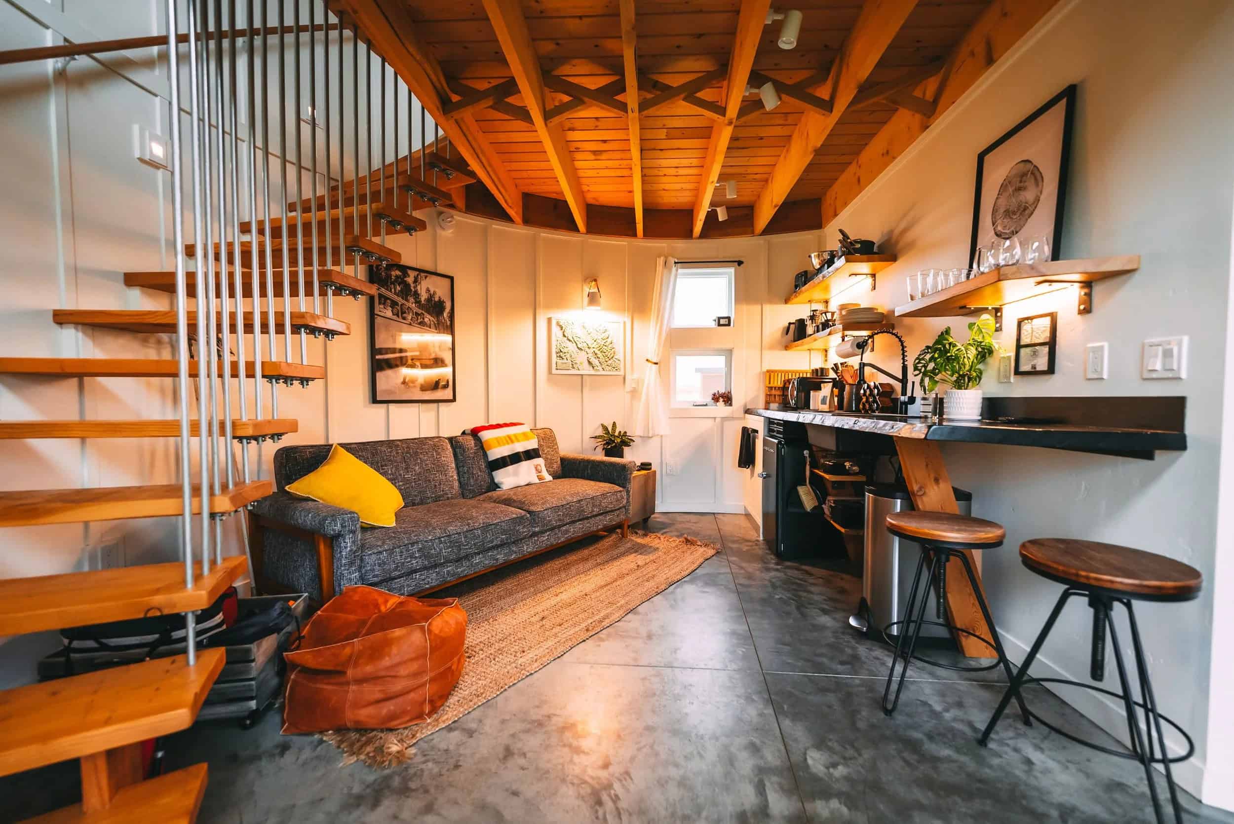 Circular silo interior with a grey sofa, tan rug, spiral wood stair, and a kitchenette along the curved white-paneled wall