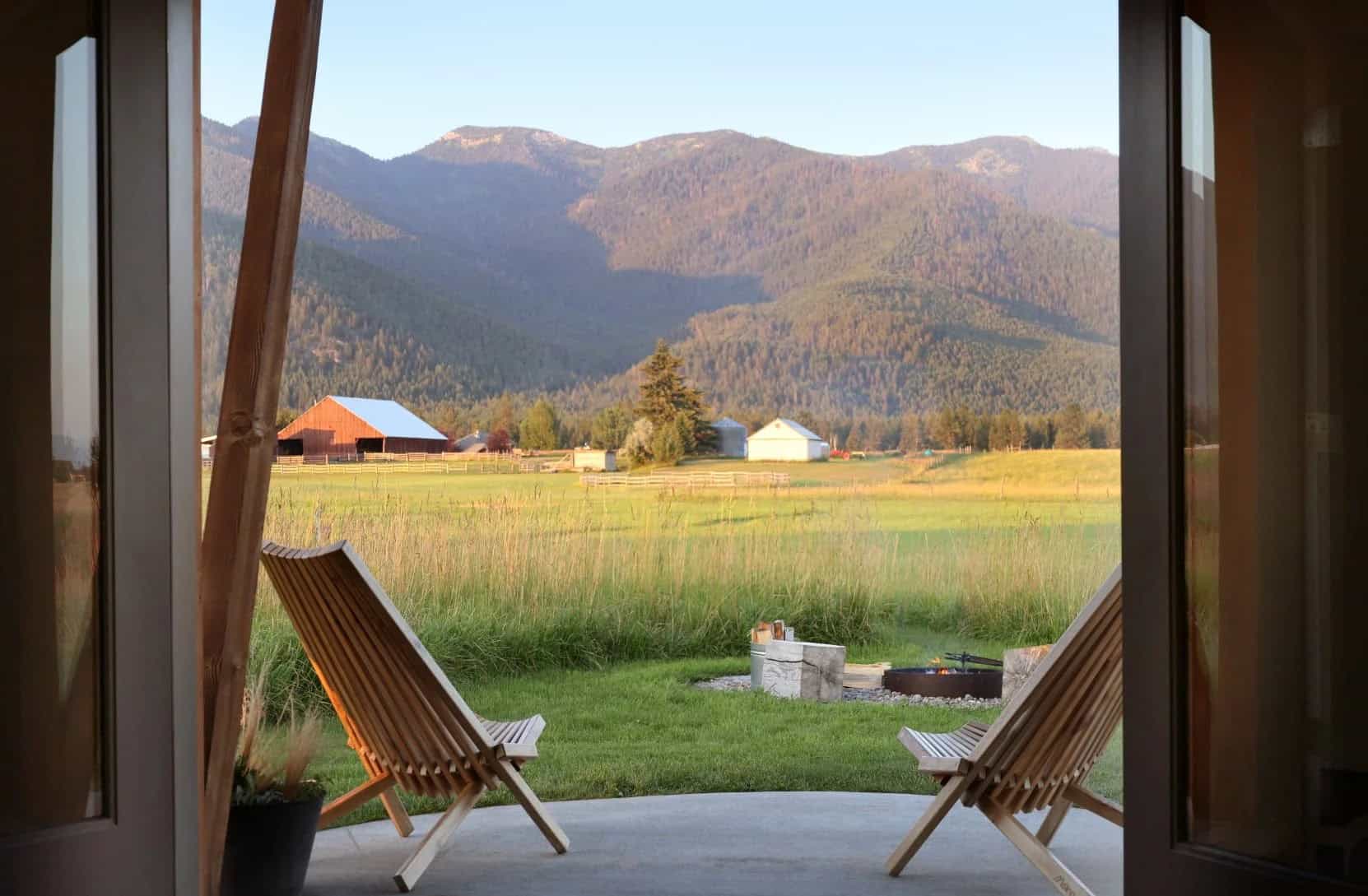 View from a silo doorway to two wood chairs on a patio, a fire pit in the grass, a red barn, and forested mountains