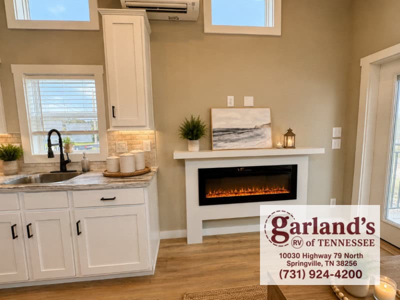 Coldwater sink under a window: marble-look counter, black gooseneck faucet, white shaker cabinets, fireplace in a white surround, mini-split above the mantel