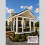 2026 Clayton Coldwater park model on a gravel lot: olive-green lap siding, white-trimmed covered porch with four square columns and black rail, dark shingled roof, clerestory windows along the side