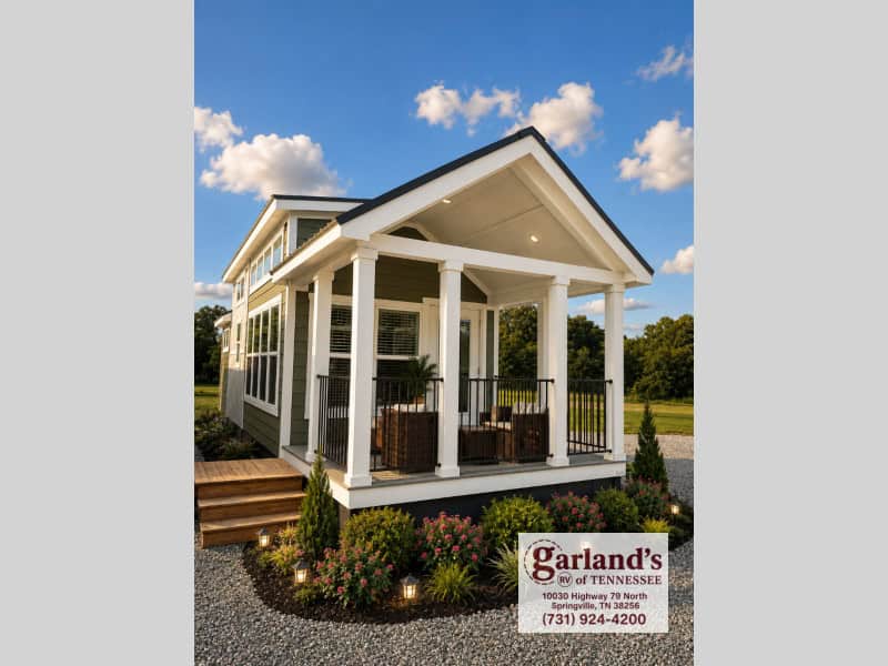 2026 Clayton Coldwater park model on a gravel lot: olive-green lap siding, white-trimmed covered porch with four square columns and black rail, dark shingled roof, clerestory windows along the side