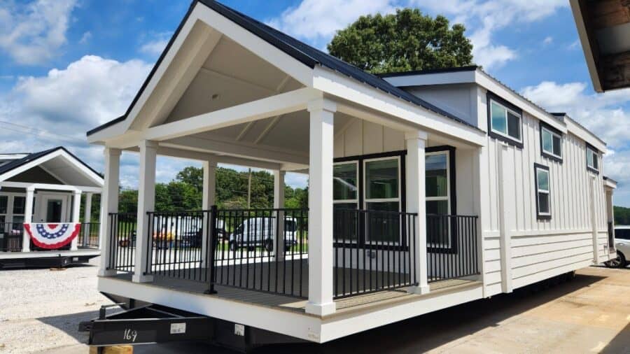 2026 Clayton Wheeler on the Springville lot: white board-and-batten, black porch rail, hitch marked 169, dark roof, gravel lot