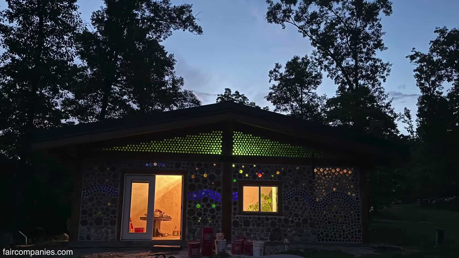 Cordwood home at dusk in the Missouri Ozarks with bands of green bottle ends glowing under the eaves, warm light in the door and window, and blue bottle patterns in the walls