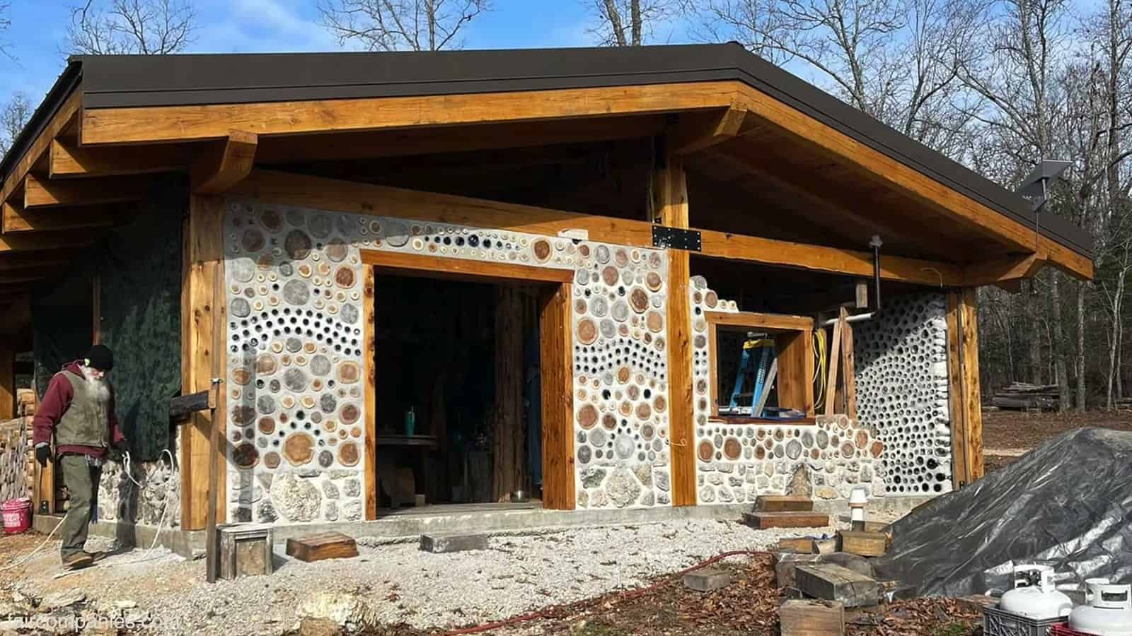 Cordwood home exterior under construction with a timber frame, deep metal roof overhang, and walls of cedar rounds and bottle mosaics
