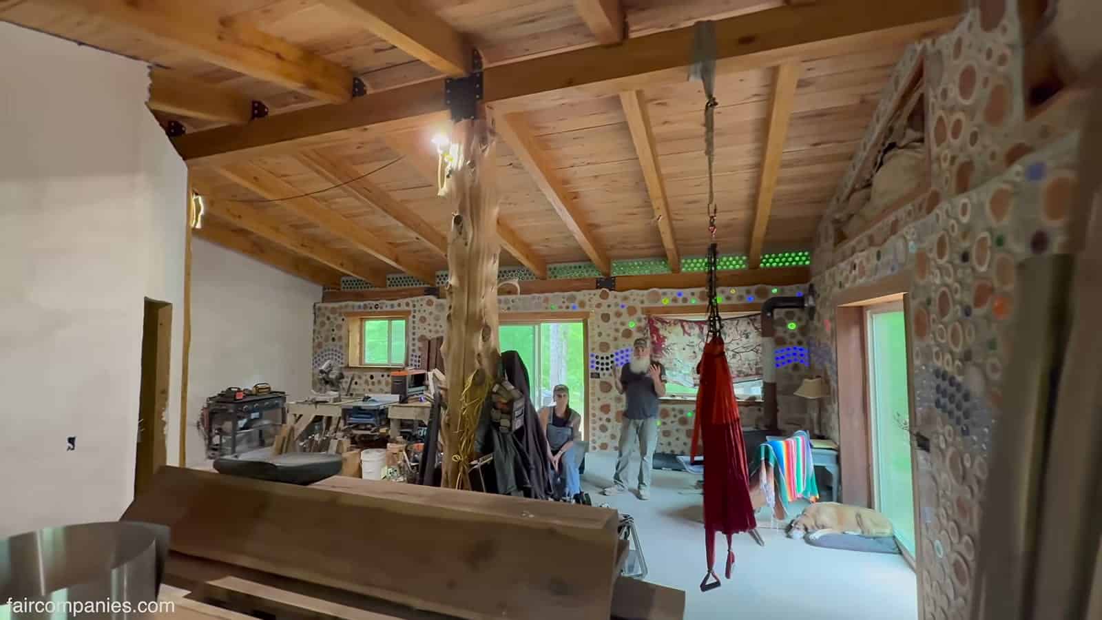 Wide interior of an unfinished cordwood home with exposed timber beams, a tree-trunk post, workshop tools, a hanging hammock, and bottle-studded walls with two people talking