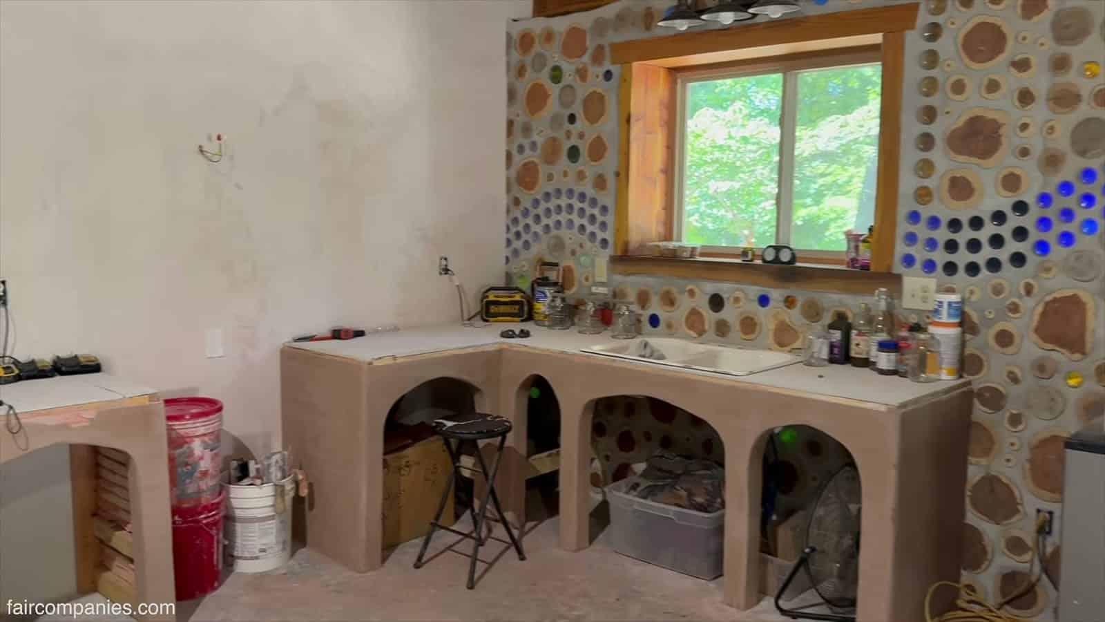 Unfinished kitchen with arched counter bases, a window over a sink, and a cordwood wall with blue glass bottles in a wave pattern